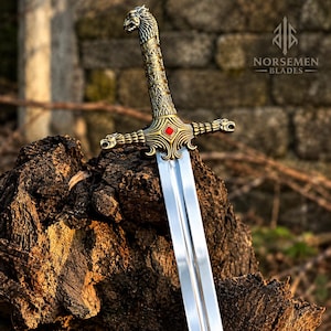 Op de afbeelding: Een metalen zwaard met een pommel in de vorm van een adelaarskop, een textuurrijke greep en een pareerstang met dierenkoppen aan de uiteinden. Een rode ruitvormige steen is in het midden van de pareerstang geplaatst. Het lemmet is zilverkleurig.
