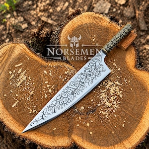 May include: A sharp kitchen knife with a pointed tip and a silver blade featuring an intricate black floral and vine engraving. The handle is dark with a detailed metallic scrollwork pattern. The text NORSEMEN BLADES is visible on the blade.
