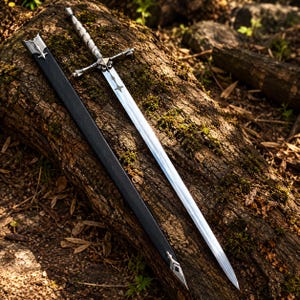 Puede incluir: Una espada de metal plateado con una guarda detallada y un mango en espiral, acompañada de una vaina negra larga con detalles metálicos plateados en los extremos, todo reposando sobre un tronco de madera texturizado y cubierto de musgo.