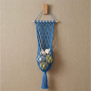 May include: A blue macrame hanging storage basket featuring a diamond-patterned net design suspended from a horizontal wooden dowel. The basket contains two white garlic bulbs and two yellow lemons, finished with a long blue tassel at the bottom.