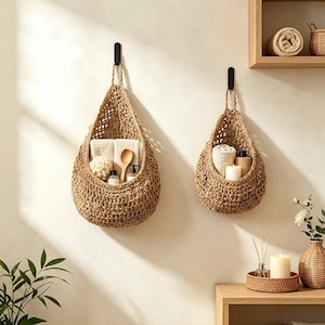 May include: Two teardrop-shaped woven baskets made of natural tan fibres, each suspended from a black wall hook. The larger basket holds folded towels, a wooden spoon, and small bottles, while the smaller basket contains a towel and a candle.
