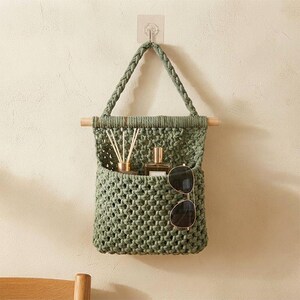 Può includere: Un organizer da parete in macramè color verde salvia con un manico intrecciato appeso a un bastoncino di legno. La tasca intrecciata contiene un diffusore di fragranze in vetro con bastoncini bianchi e occhiali da sole con montatura sottile.