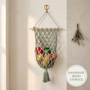 Puede incluir: Una cesta colgante para frutas de macramé en color verde salvia con un diseño de red anudada suspendida de un palo de madera. La cesta contiene varias frutas, incluyendo manzanas rojas, una lima verde y limones amarillos, con una borla decorativa.