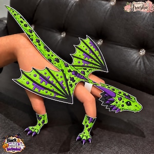 May include: A flat, paper-like dragon costume accessory featuring a vibrant lime green and purple slime-drip pattern. The design includes a long tail, two wings, a head piece with purple spikes, and two clawed foot covers, all with black outlines.