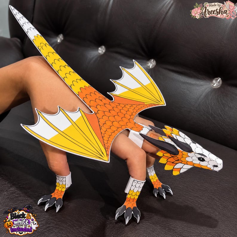 May include: A paper dragon finger puppet featuring a candy corn colour scheme of orange, yellow, and white. The dragon has detailed scales, pointed wings, and sharp claws. The design includes a long tail and a head with black horns.