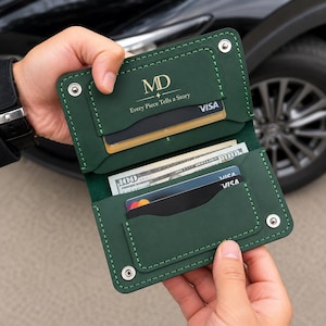 以下が含まれることがあります： 銀色の金属製スナップボタン、目に見えるステッチ、複数のカードスロットを備えたダークグリーンのレザーウォレット。財布には「MD」の文字と「Every Piece Tells a Story」というテキストが刻印されており、数枚のクレジットカードと現金が入っています。