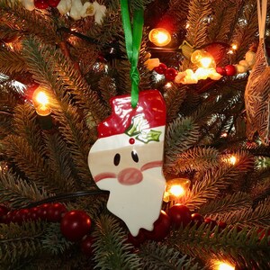 May include: A white ceramic Christmas ornament shaped like Santa Claus's head with a red hat and white beard. The ornament is hanging on a Christmas tree with red berries, white flowers, and lights.