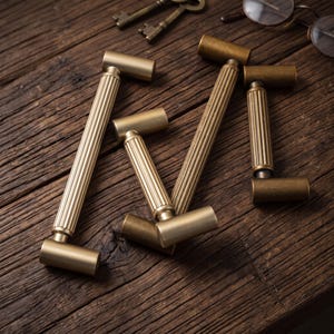 May include: A collection of four brass cabinet pulls featuring a cylindrical design with a central fluted, ribbed texture and smooth, solid end caps. The hardware has a matte, brushed metallic finish and is arranged in a scattered, overlapping pattern.