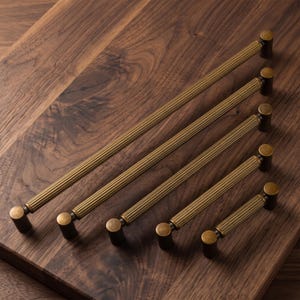 May include: Five brass cabinet pulls of varying lengths arranged diagonally. Each pull features a cylindrical, ribbed handle with a metallic gold finish and dark, rounded end caps. The design is consistent across all five pieces, showcasing a textured, grooved surface on the main bar and smooth, solid circular mounts at both ends.