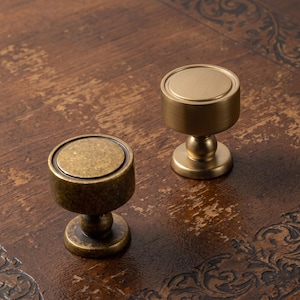 May include: Two round cabinet knobs with a cylindrical body and a pedestal base. One knob features a smooth, brushed brass finish, while the other has a textured, antique bronze finish with visible dark spots and a weathered appearance.