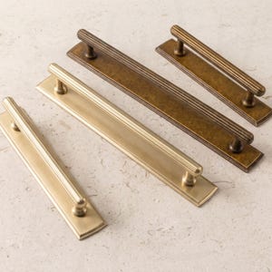 May include: Three metal drawer pulls with a long rectangular backplate and a ribbed cylindrical handle. The pulls are arranged diagonally, featuring two in a brushed brass finish and one in a darker, aged bronze finish with visible texture.