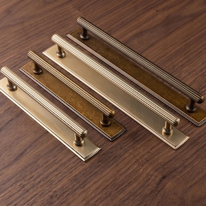 May include: A set of three metal drawer pulls with a fluted, cylindrical handle design. Each pull is mounted on a rectangular backplate. The set includes two pulls with a brushed brass finish and one pull with a dark, antique bronze finish.