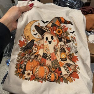 Może przedstawiać: Kremowy t-shirt z nadrukiem graficznym przedstawiającym ducha w kapeluszu czarownicy i płaszczu w stylu patchwork. Duch trzyma zapaloną latarnię, otoczony dyniami, czarnym krukiem, jesiennymi liśćmi, półksiężycem i nietoperzami na ciemnym tle.