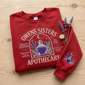 May include: A red crewneck sweatshirt featuring embroidered text reading Owens Sisters Apothecary, a central potion bottle graphic, and floral accents. The design includes quotes about salt and rosemary, with a matching potion motif on the sleeve.