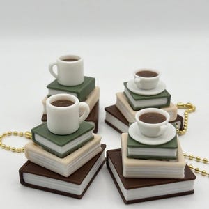 May include: Four miniature keychains featuring stacks of three books in shades of brown, beige, and green. Each stack is topped with a small white ceramic-style mug filled with a dark brown liquid, some with a saucer, attached to a gold ball chain.