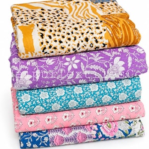May include: A stack of five folded cotton fabrics. The top piece features a yellow and black animal print. Below it is a purple floral fabric with the text JaipurTextilesCo, followed by teal, pink, and blue floral patterned textiles.