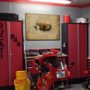 May include: A vintage illustration of a yellow motorcycle on a brown background. The motorcycle has a black seat and black wheels. The illustration is framed and hanging on a wall in a garage.