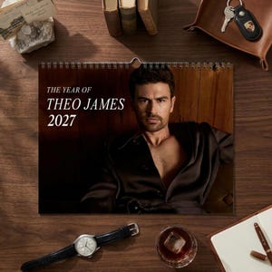 Puede incluir: Un calendario de pared titulado THE YEAR OF THEO JAMES 2027 que presenta el retrato de un hombre con cabello oscuro y barba, vistiendo una camisa marrón oscuro de cuello abierto sobre un fondo de paneles de madera, con espiral metálica plateada.