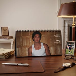 May include: A 2027 spiral-bound desk calendar featuring a portrait of Belmont Cameli wearing a white tank top against a wood-paneled background. The text on the calendar reads Belmont Cameli 2027 Calendar. The calendar is displayed on a wooden desk.