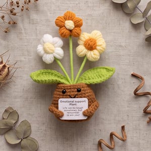 May include: A crocheted brown pot with a smiling face holding a white sign that reads Emotional support Plant Life can be tough sometimes, but remember to are you! Three crocheted flowers in orange, white, and yellow with green leaves.