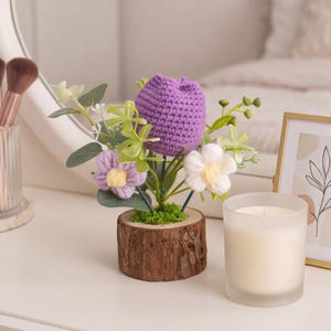 Op de afbeelding: Een decoratief arrangement met een grote gehaakte paarse tulp, een kleinere lichtpaarse gehaakte bloem en een witte gehaakte bloem met een geel hart, alles geplaatst in een natuurlijke houten voet met groen mos en kunstgroen.