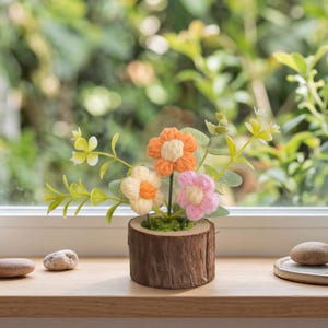 Op de afbeelding: Een decoratief arrangement met drie kleine, gehaakte bloemen in oranje, crème en lichtroze, geplaatst in een plantenbak van een natuurlijke houten schijf. De bloemen zijn geaccentueerd met groen kunstblad en mos voor een compact geheel.