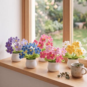 May include: Five small white ceramic pots each containing a crocheted flower arrangement. The flowers feature purple, blue, pink, and yellow petals with green crocheted leaves. A speckled ceramic mug sits to the right of the flower pots.