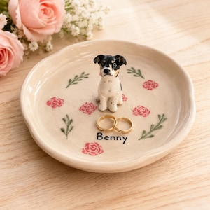 May include: A round, cream-coloured ceramic trinket dish featuring a hand-painted black, white, and tan dog figurine in the centre. The dish is decorated with small pink rose motifs and green sprigs, with the name Benny printed in black text.