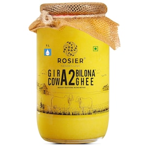 Op de afbeelding: Een glazen pot van 1L met gele ghee en een deksel bedekt met jute. Het etiket bevat de tekst ROSIER NATURE'S LOVE GIR COW A2 BILONA GHEE, een afbeelding van twee koeien in een veld en een groen vierkant icoon voor vegetarische producten.