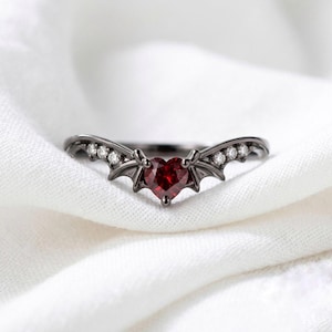 Puede incluir: Un anillo de metal negro con una piedra preciosa roja en forma de corazón en el centro, flanqueada por motivos de alas de murciélago a cada lado. La banda está decorada con tres pequeñas piedras claras de corte redondo en cada ala.