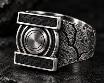 Puede incluir: Un anillo de tono plateado con un emblema circular central de anillos concéntricos, flanqueado por dos barras rectangulares horizontales con un acabado oscuro y texturizado. La banda tiene una superficie rugosa y desgastada con una grieta.