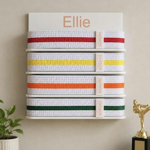 Op de afbeelding: Een wit wandrek voor vechtsportbanden met de naam Ellie bovenaan gedrukt. Het rek bevat vier witte banden met rode, gele, oranje en groene strepen, elk vastgezet met een label met de datums: 14/09/25, 21/12/25, 29/03/26 en 29/06/26.