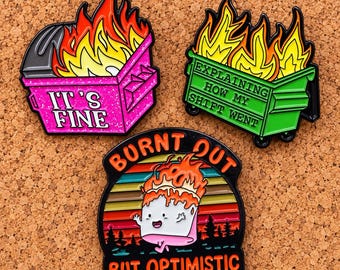 May include: Three enamel pins featuring a pink dumpster with flames and the text IT'S FINE, a green dumpster with flames and the text EXPLAINING HOW MY SHIFT WENT, and a marshmallow character on fire with the text BURNT OUT BUT OPTIMISTIC.