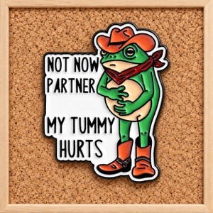 Könnte beinhalten: Eine Metall-Emaille-Anstecknadel mit einem grünen Frosch, der einen orangefarbenen Cowboyhut, ein rotes Halstuch und orangefarbene Cowboystiefel trägt. Der Frosch hält seinen Bauch mit beiden Händen. Der weiße Hintergrund enthält den schwarzen Text: NOT NOW PARTNER MY TUMMY HURTS.