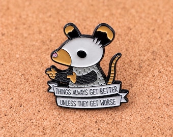 Puede incluir: Un pin de esmalte metálico que muestra un oposum de dibujos animados con cara blanca, orejas negras y hocico color canela. El oposum gesticula con sus patas sobre una pancarta blanca con el texto negro: THINGS ALWAYS GET BETTER UNLESS THEY GET WORSE.