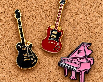 Puede incluir: Tres pines de esmalte con forma de instrumentos musicales: una guitarra eléctrica negra con bordes dorados, una guitarra eléctrica roja con bordes dorados y un piano de cola rosa con detalles negros en las teclas y la tapa.