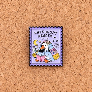 Könnte beinhalten: Ein rechteckiger Emaille-Pin in Form einer Briefmarke mit gewelltem schwarzem Rand. Auf hellviolettem Grund ist eine weiße Ente unter einer geblümten Decke zu sehen, die mit einer Taschenlampe ein rosa Buch liest. Text LATE NIGHT READER CLUB.