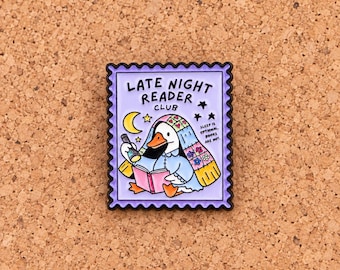 Puede incluir: Un pin de esmalte rectangular con forma de sello postal y borde negro festoneado. El fondo morado claro muestra un pato blanco bajo una colcha floral, leyendo un libro rosa con una linterna. El texto dice LATE NIGHT READER CLUB.
