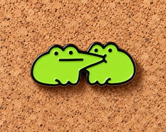 Puede incluir: Un broche de esmalte verde lima brillante que muestra dos ranas estilizadas. La rana de la izquierda tiene un hocico largo y puntiagudo que se extiende hacia la rana de la derecha, que tiene una cara redondeada y ojos de punto negro.