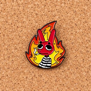 May include: A metal enamel pin featuring a red rabbit character with large black eyes, wearing a black and white striped shirt, holding a lit cigarette, and positioned in front of a bright yellow and orange flame design with a black outline.