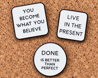 Puede incluir: Tres pines de esmalte blanco con bordes y texto en negro. Un pin cuadrado dice YOU BECOME WHAT YOU BELIEVE, otro cuadrado dice LIVE IN THE PRESENT y un pin circular tiene el texto DONE IS BETTER THAN PERFECT.