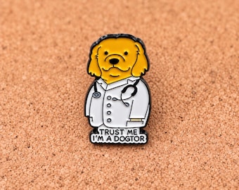 Puede incluir: Un pin de esmalte dorado y blanco que muestra un perro de dibujos animados con orejas caídas, vistiendo una bata de médico blanca y un estetoscopio negro al cuello. El texto en la parte inferior dice "TRUST ME I'M A DOGTOR" en letras negras.