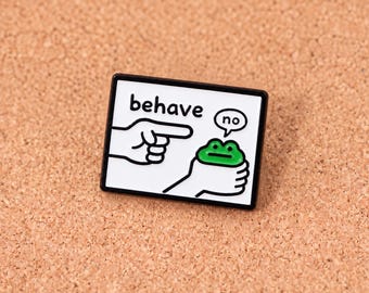 Puede incluir: Un pin de esmalte rectangular con fondo blanco y borde negro. Presenta un dibujo de línea negra de una mano señalando a una pequeña rana verde sostenida por otra mano. El texto "behave" está impreso en negro y un globo de texto dice "no".