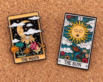 Puede incluir: Dos pines rectangulares de esmalte con diseños de cartas de tarot. El pin izquierdo muestra una luna creciente, nubes y flores coloridas con el texto THE MOON. El pin derecho muestra un sol, nubes y flores con el número romano X y THE SUN.