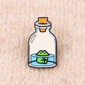 May include: A metal enamel pin featuring a clear glass bottle with a cork stopper. Inside the bottle, a small green frog sits in a pool of light blue water. The design has a black outline and a smooth, glossy finish, capturing the frog resting.