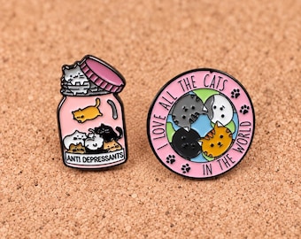 Puede incluir: Dos pines de esmalte. Uno es un frasco rosa lleno de gatos pequeños con el texto ANTI DEPRESSANTS. El otro es un pin circular que muestra un globo terráqueo con caras de gatos y el texto I LOVE ALL THE CATS IN THE WORLD con huellas negras.