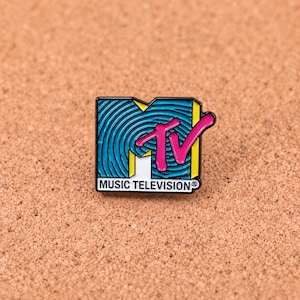 Könnte beinhalten: Eine rechteckige Emaille-Anstecknadel mit dem MTV-Logo, einem blauen Hintergrund mit konzentrischen kreisförmigen Linien, einem leuchtend rosa TV-Schriftzug, gelben Akzenten und dem Text MUSIC TELEVISION in Schwarz auf weißem Grund.