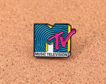 Puede incluir: Un pin de esmalte rectangular con el logotipo de MTV, un fondo azul con líneas circulares concéntricas, letras TV en rosa brillante, detalles amarillos y el texto MUSIC TELEVISION impreso en negro sobre una base de color blanco.