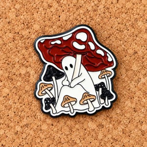 May include: A metal enamel pin featuring a white ghost figure standing under a large red mushroom cap with white spots. The design includes several smaller mushrooms in black, white, and tan colors surrounding the central ghost figure.