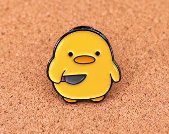 Könnte beinhalten: Ein gelber Emaille-Pin in Form eines runden Kükens mit schwarzen Augen und einem orangefarbenen Schnabel. Das Küken hält ein kleines graues Messer mit einem rosa Griff in seinem Flügel. Das Design hat kräftige schwarze Konturen und eine glatte Oberfläche.